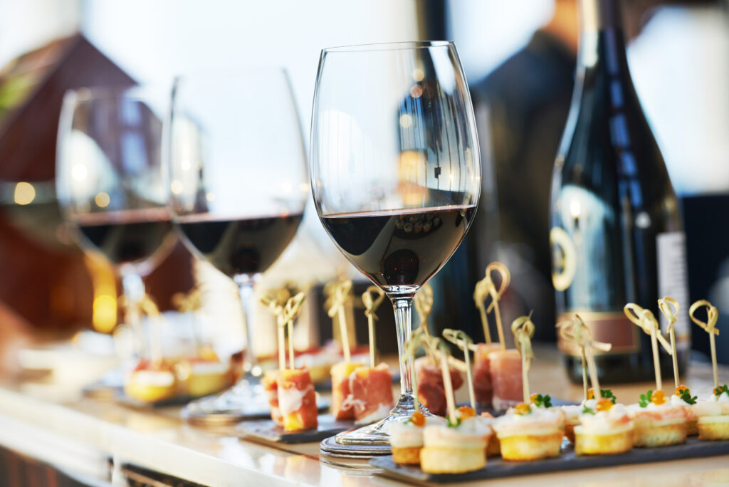 Glasses of a variety of red wines lined up neatly alongside several types of hors d’oeuvres.
