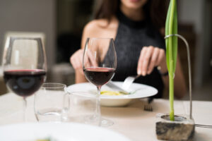 Glass of wine and small green plant in front of someone eating from a plate with a fork.