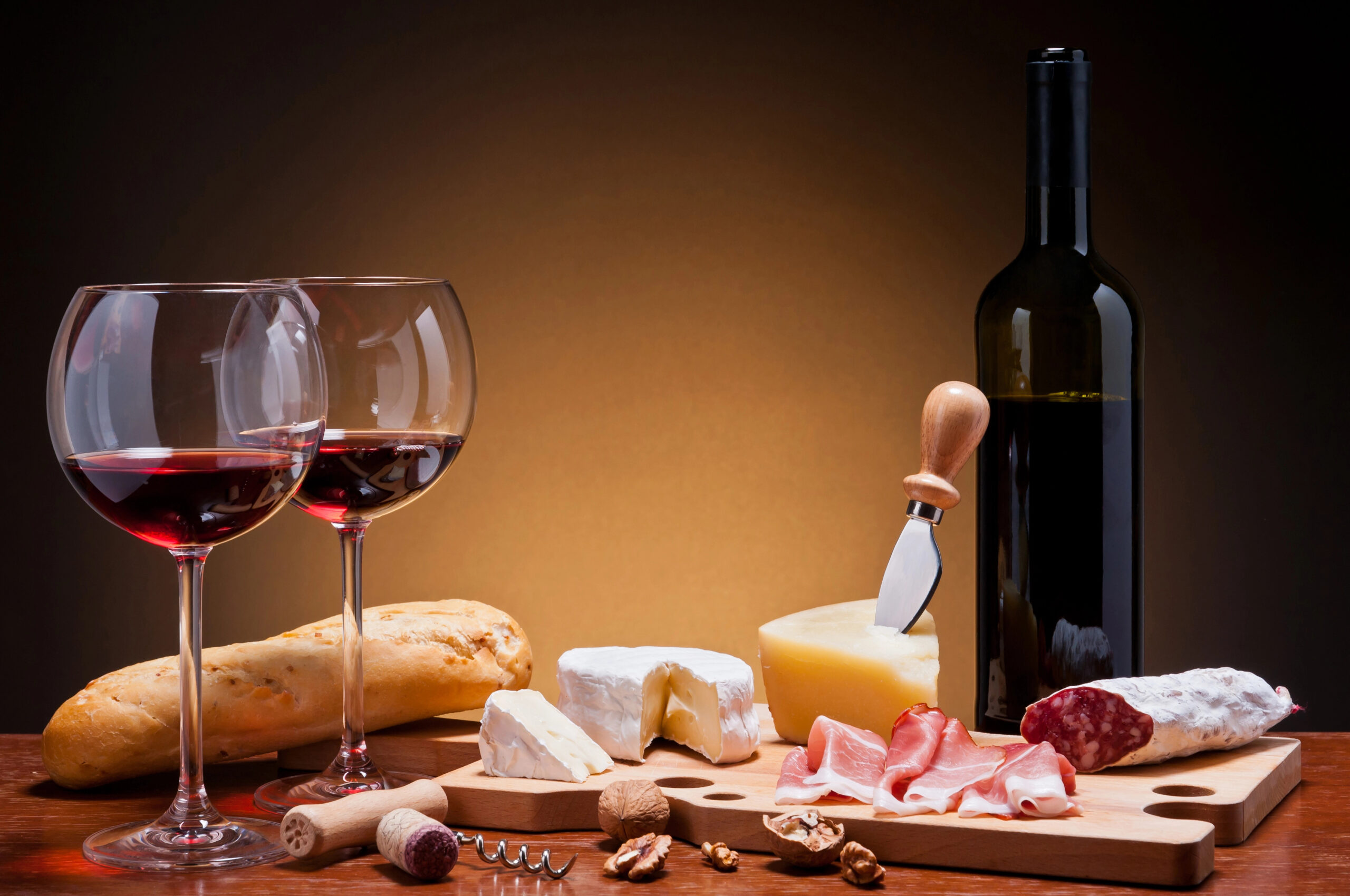 Bottle and glasses of red wine, corkscrews, and a cutting board with charcuterie, cheeses, and bread.