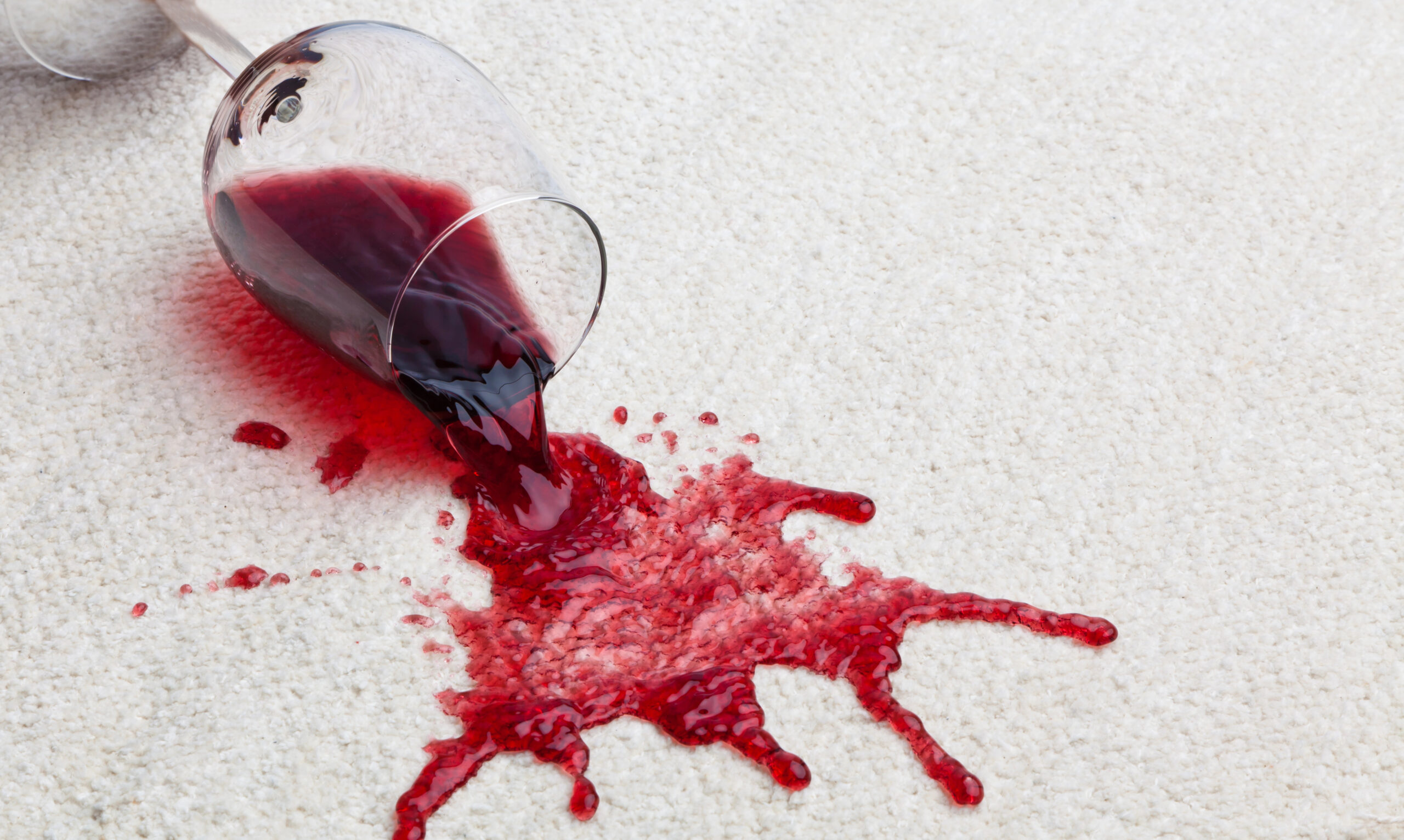 A glass of red wine spilling onto white carpet.