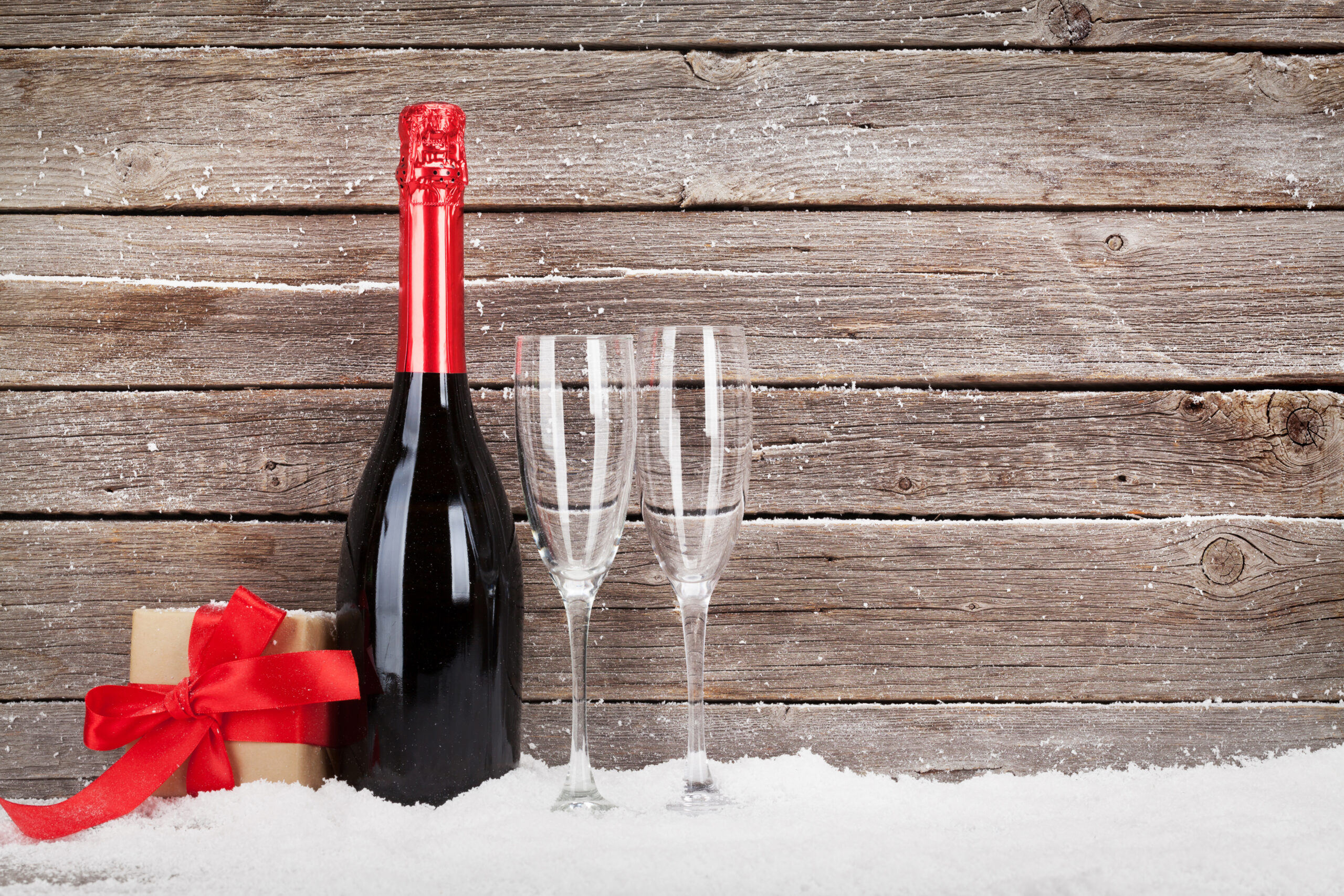 Bottle of red wine in the snow, next to two empty glasses and a small wrapped gift box.